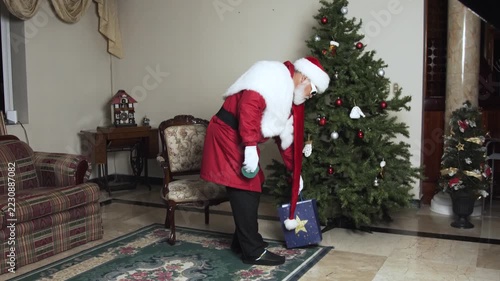 Modern Santa walks past Christmas tree after leaving blue gift box