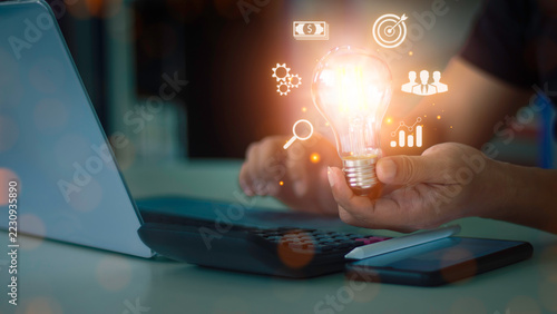 Businessman holding glowing light bulb with virtual business icon overlay for creative idea, startup strategy, and smart business solution concept.