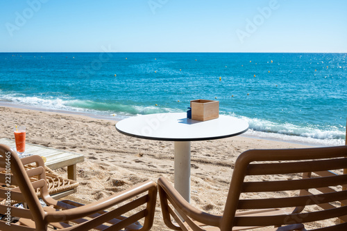 Beach table and chairs on the sand beside calm ocean on sunny day.