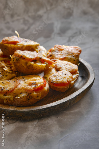 Roasted potatoes topped with tomato, melted cheese and herbs on wooden platter. Baked potato dish for a savory meal. Comfort food and home cooking. Vegetarian side dish with copy space.