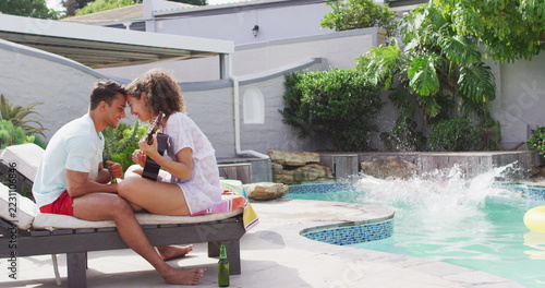 Happy diverse couple in love playing guitar, their friend jumping into swimming pool at pool party