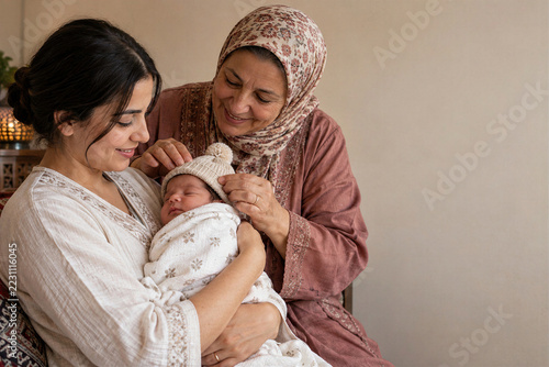 Young middle eastern mother holding newborn baby in knitted hat with grandmother in headscarf smiling gently at home