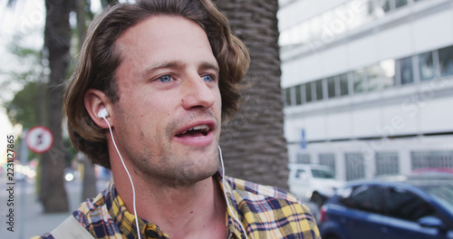 A man in a checkered shirt enjoys music on his phone while exploring the city.
