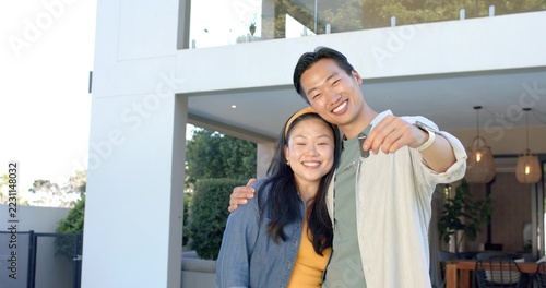 Asian couple smiling at modern 2-story house entry, man holding keys, woman wearing denim jacket