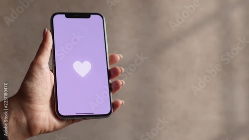 A hand holds a smartphone with a purple screen where a white heart icon with waves pulses and fades, illustrating a digital like or notification in a minimalist setting.