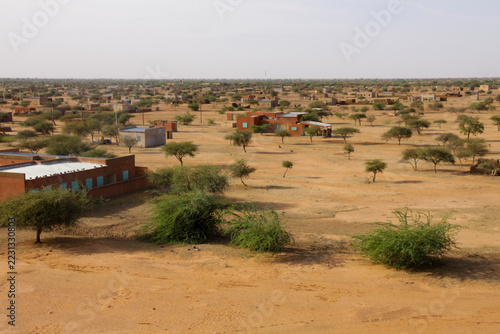 Person walking across a sandy plain with acacia trees and buildings, Sahel Region, Markoye, Burkina Faso
