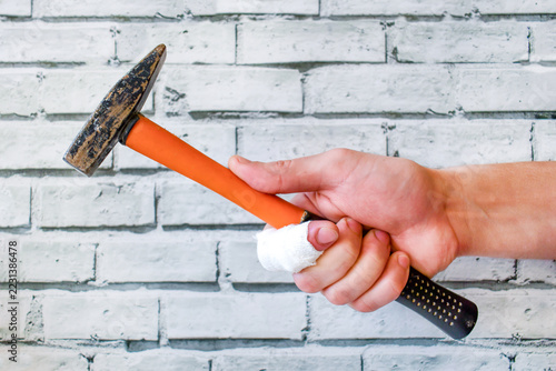 Hand with bandaged finger holding hammer with orange handle against white brick wall. Work injury and DIY accident concept for occupational safety and insurance industry.