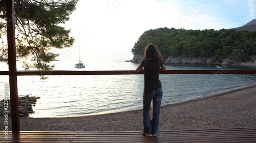 Woman standing on wooden terrace at sunset looking over calm bay