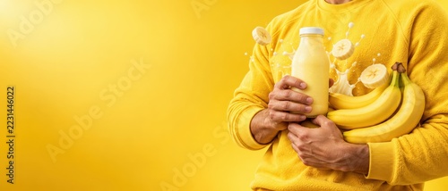 Person in yellow sweater holding banana milk bottle and fresh bananas with milk splash on yellow background with copy space