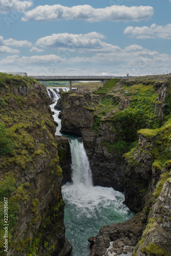 Barnafoss waterfall rushes rapidly through a rugged volcanic gorge below a pedestrian footbridge in western Iceland under a cloudy sky.