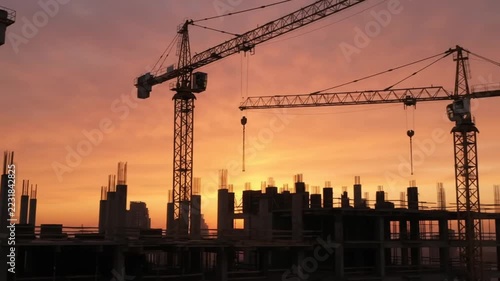 Construction cranes and unfinished building silhouetted against an orange sunset sky with tranquil mood