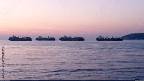 Cargo ships anchored on calm open water at sunset under a pink twilight sky