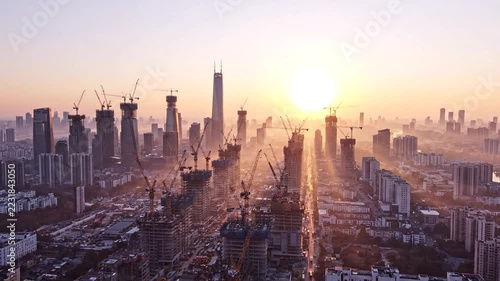Skyscraper construction cranes over a dense city skyline at sunrise, aerial timelapse with soft haze and warm backlight