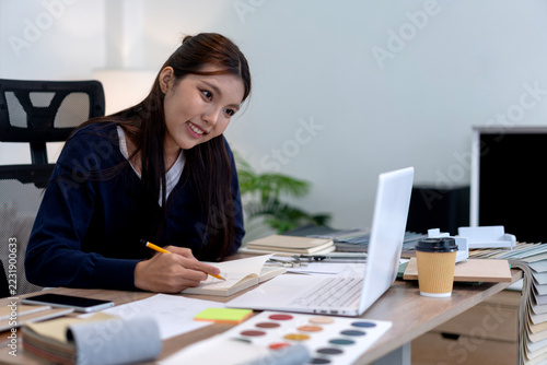 Young Asian Female Furniture Designer Working on Creative Sketches at Modern Office Desk with Laptop