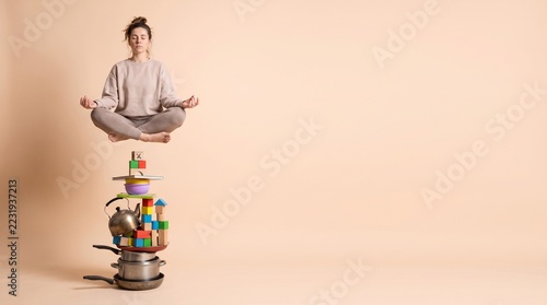 Woman meditating in lotus position floating above a stack of household items on beige background with space for text