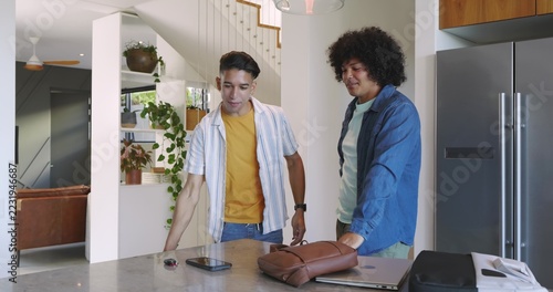 Diverse male friends leaning on island in kitchen, holding smartphone, keys, leather bag and laptop