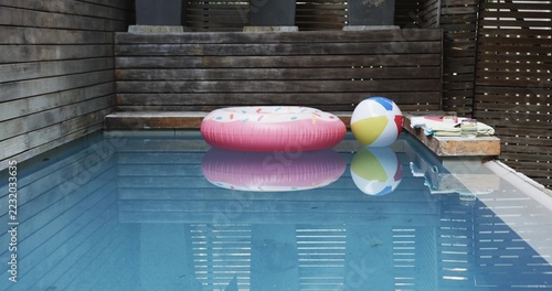 Pink donut float and multicolored beach ball are floating in rectangular pool near wooden ledge