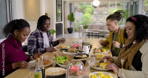 Diverse friends sitting around wooden dining table sharing salad, passing plates, holding glasses