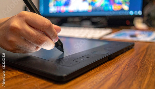 Digital Art Creation: A close-up shot of a hand using a stylus on a digital drawing tablet, with a computer monitor displaying colorful artwork in the background, signifying the creative process.