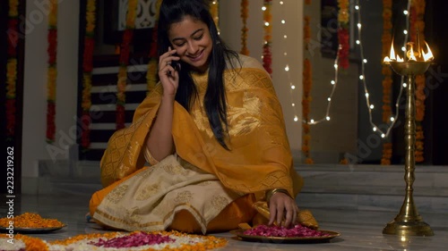 Diwali celebration and decoration - Pretty Indian girl in traditional dress speaking over the phone and making flower rangoli . 