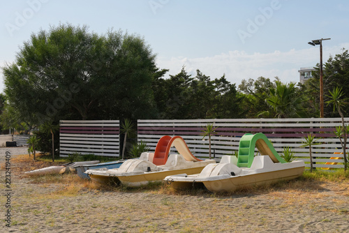 Two old pedal boats with orange and green water slides and kayaks resting on sandy beach by wooden fence at end of season, Calabria, Italy