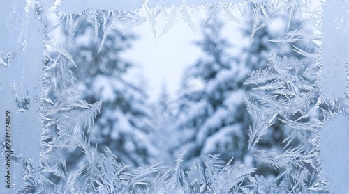Frost patterns on window glass framing blurred snowy pine trees in winter season