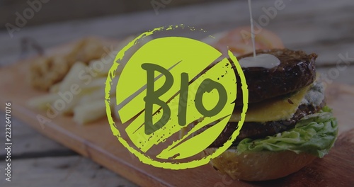 Pouring sauce over stacked burger on wooden board at rustic table, with fries and BIO graphic