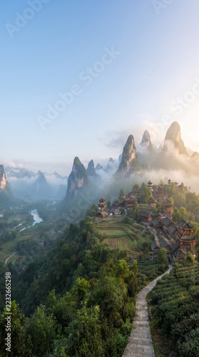 Scenic mountain village nestled among misty karst peaks with a narrow path leading through lush green fields at sunrise