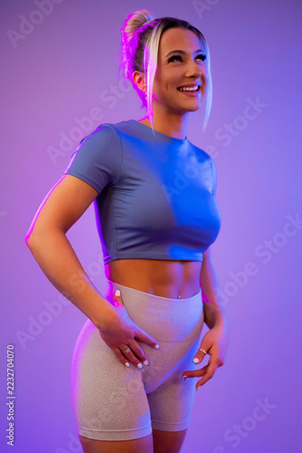 Smiling athletic woman in workout clothes posing under neon purple and blue lighting