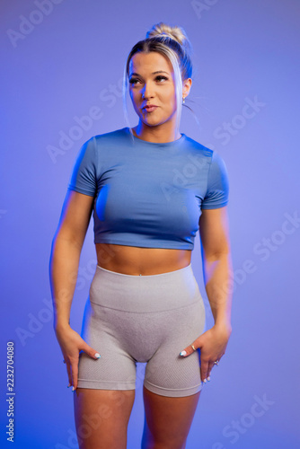 Fit young woman in blue crop top and gray leggings posing confidently against purple background