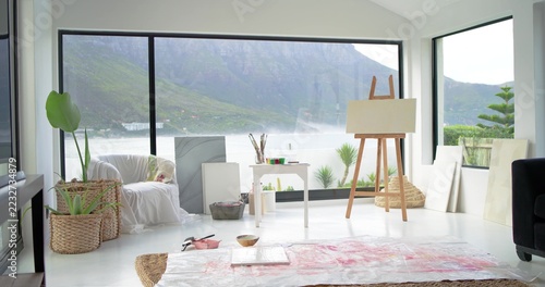 Easel holding blank canvas near paint table with brushes, drop cloth, windows showing misty lake