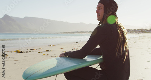 Obraz na plátně Image of smiling caucasian man with dreadlocks in wetsuit sitting with surfboard