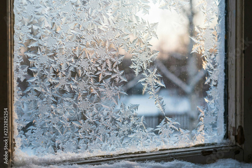 Fotografie Intricate frost patterns on a window pane with a blurred winter landscape visibl