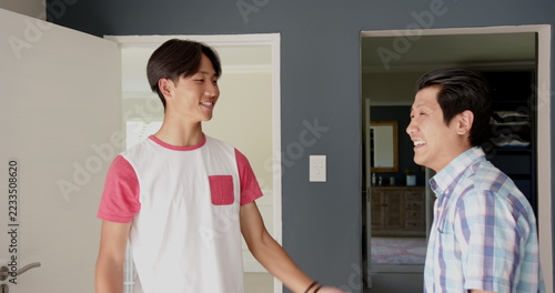 Asian father and teenage son smiling and greeting each other by white door in home entryway