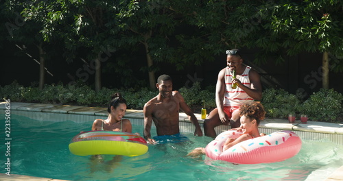 Diverse friends lounging on donut floats sipping from bottles and wearing sunglasses in backyard