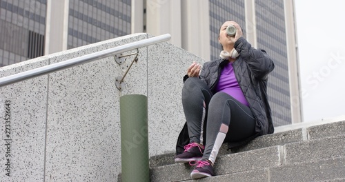 Senior woman sitting on steps in plaza drinking from bottle, wearing jacket, copy space