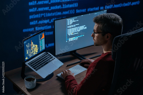 Concentrated male coder works at multi-monitor setup in a dimly lit room with lines of code projected in the background. Software engineer tackles a challenging machine learning script late at night