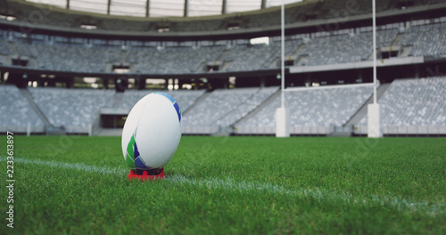 Rugby ball on tee on rugby pitch at empty sports stadium with tiered seating, copy space