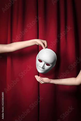 Obraz na plátně A white mask hangs between two hands against a deep red curtain, as one hand holds it above the open palm of the other