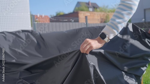 Woman covering outdoor sofa with protective furniture cover on patio. Pulling fabric over cushions to protect garden furniture from weather