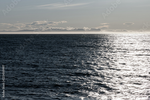 Sunlight shimmers across rippled water in Hunafjordur, North Iceland, with a misty distant coastline beneath pale clouds. Muted colors and deep shadows evoke a vintage film look.