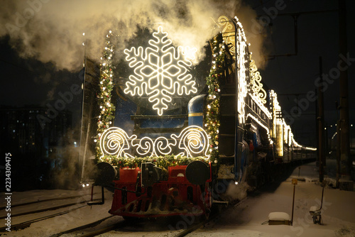 Decorated Christmas steam locomotive travels on snowy railway during frosty winter morning. Illuminated snowflake, garland and festive lights cover retro engine pulling holiday passenger carriage.