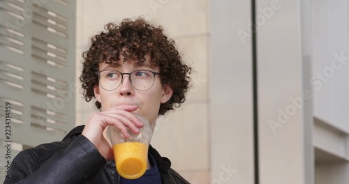 Man standing near metal vent panel, sipping orange juice, in leather jacket and eyeglasses