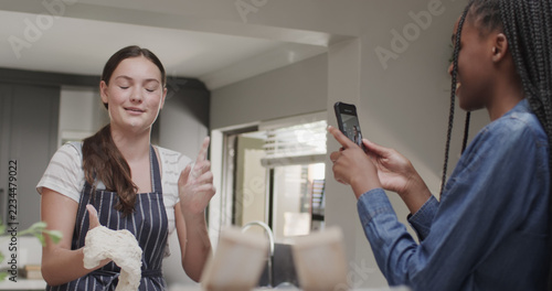 Happy diverse girls preparing meal and taking photos with smartphone in kitchen at home