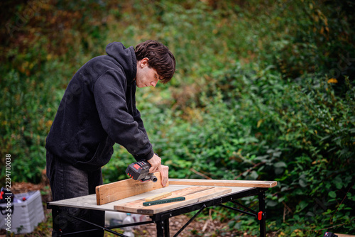Fotografie Male woodworker using a battery-powered sander on a wooden plank clamped to a folding table outdoors