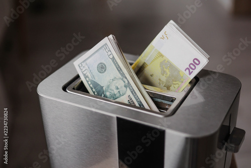 US dollar bills and euro banknotes sticking out of a toaster, symbolizing money-making, financial risk, inflation, currency exchange, and the pressure to generate income.