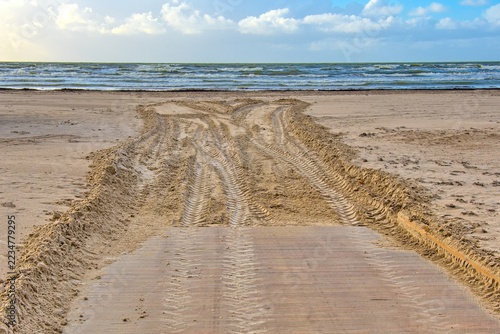 Wooden boardwalk path ending at a sandy sea beach with deep tractor tire tracks. Baltic sea waves and blue sky with clouds on the horizon.