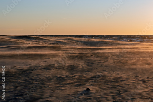 Wind blowing sand across coastal dunes on a sandy beach during an early August sunset with warm side light from the right.