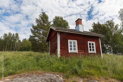 Small red wooden cabin with white window frames on a grassy summer meadow.