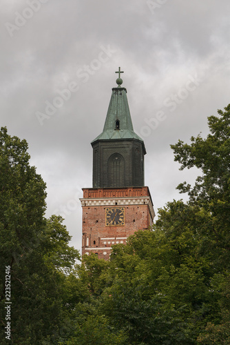 Turku Cathedral tower rises above dense green summer trees under a gray sky.